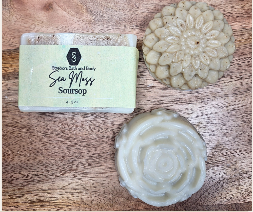 Three bars of organic sea moss and soursop soap on a wooden surface, with one bar packaged in a cardboard box labeled 'Strebors Sea Moss Soap', and the other two displayed unpackaged.